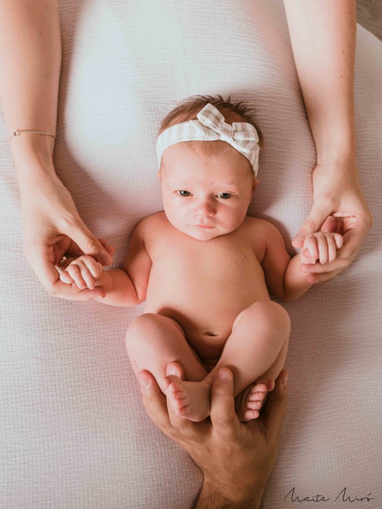 Fotografía de bebé sujetada dulcemente por las manos por su madre y su padre sosteniéndole los pies
